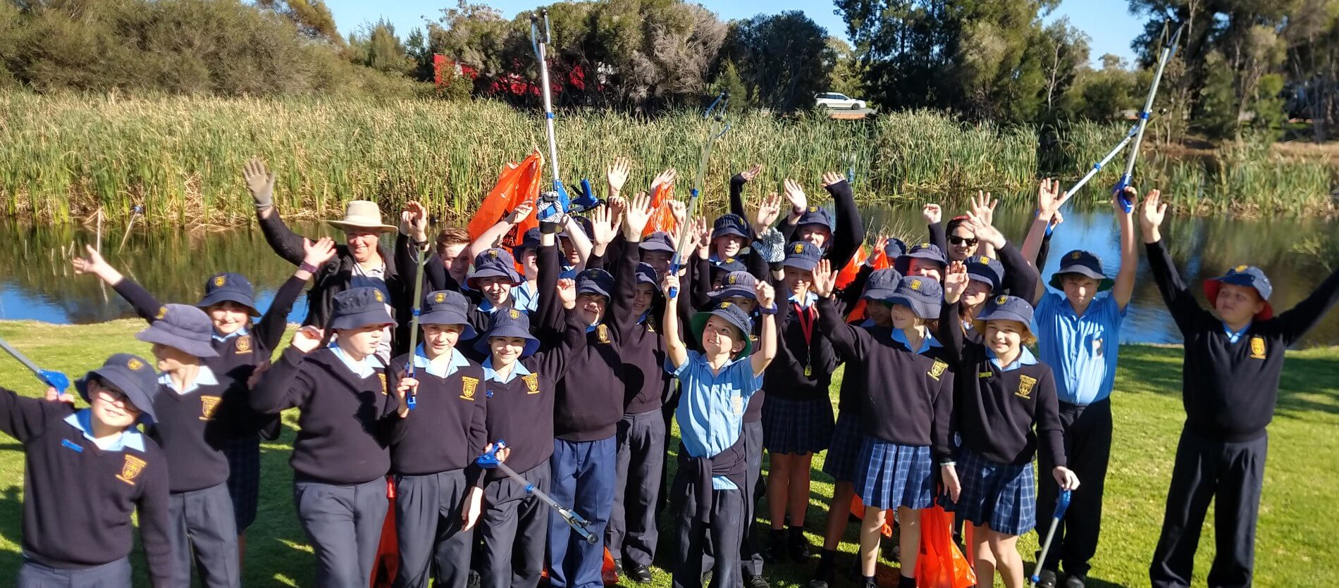 AlargegroupoflocalprimaryschoolstudentsholdingrubbishpickersandrubbishbagscelebratingtheireffortscollectingrubbisharoundalocallakelocatedattheOllieWorrellReserve