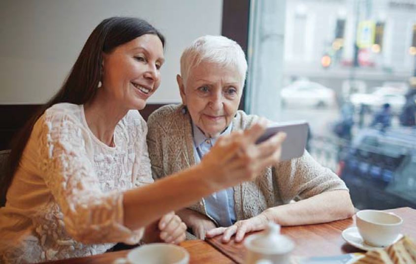 A lady holding her phone in front of an older lady near a window in a cafe.