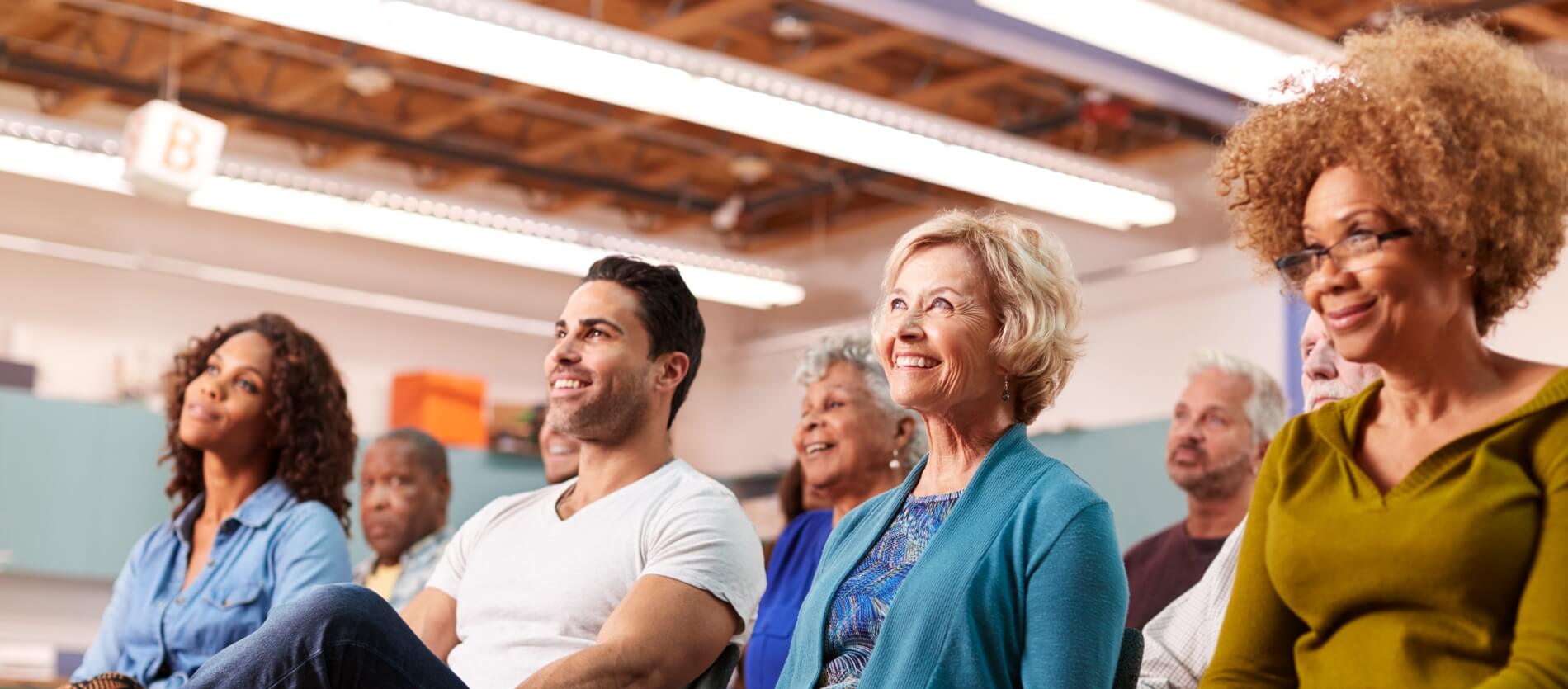 A community group sitting in a larger community hall listening and/or watching something happening in front of them.