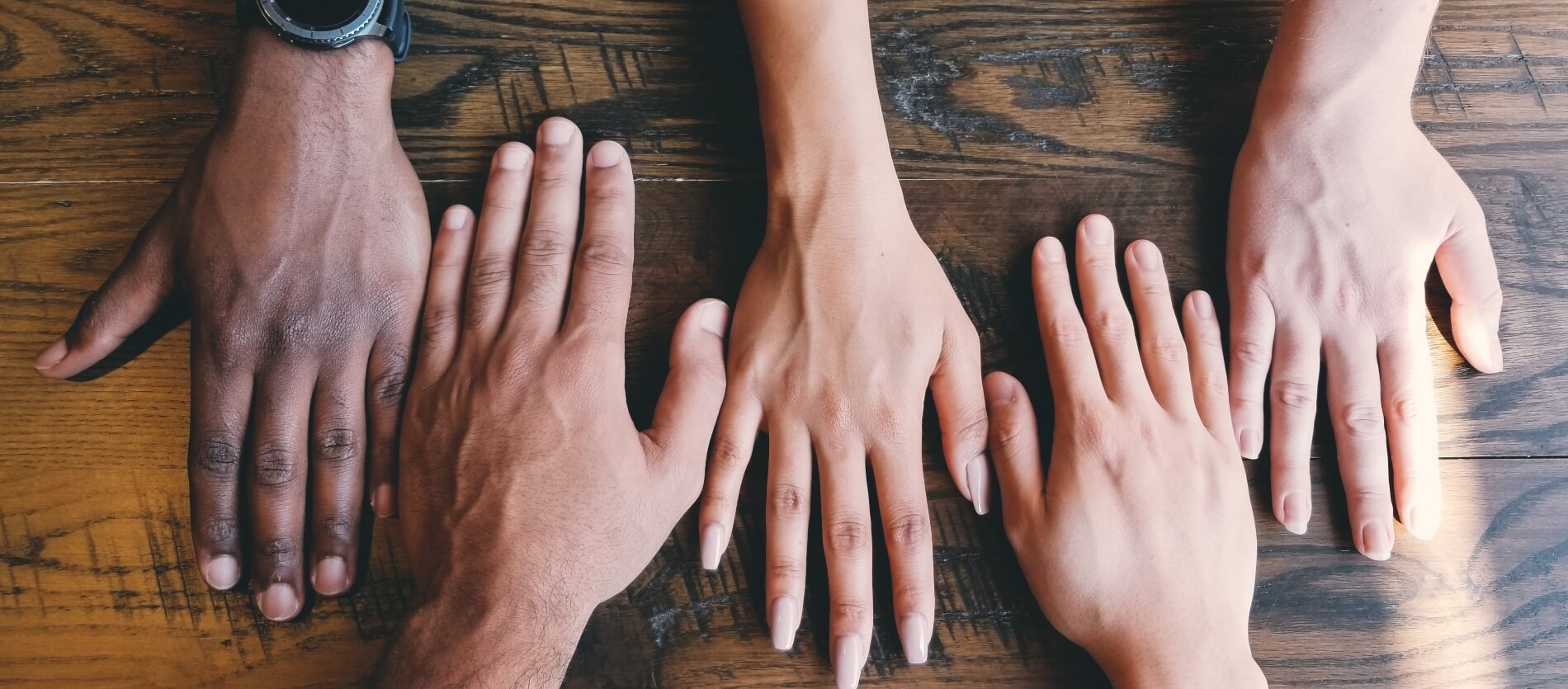 Five differing hands palm down on a wooden brown table.