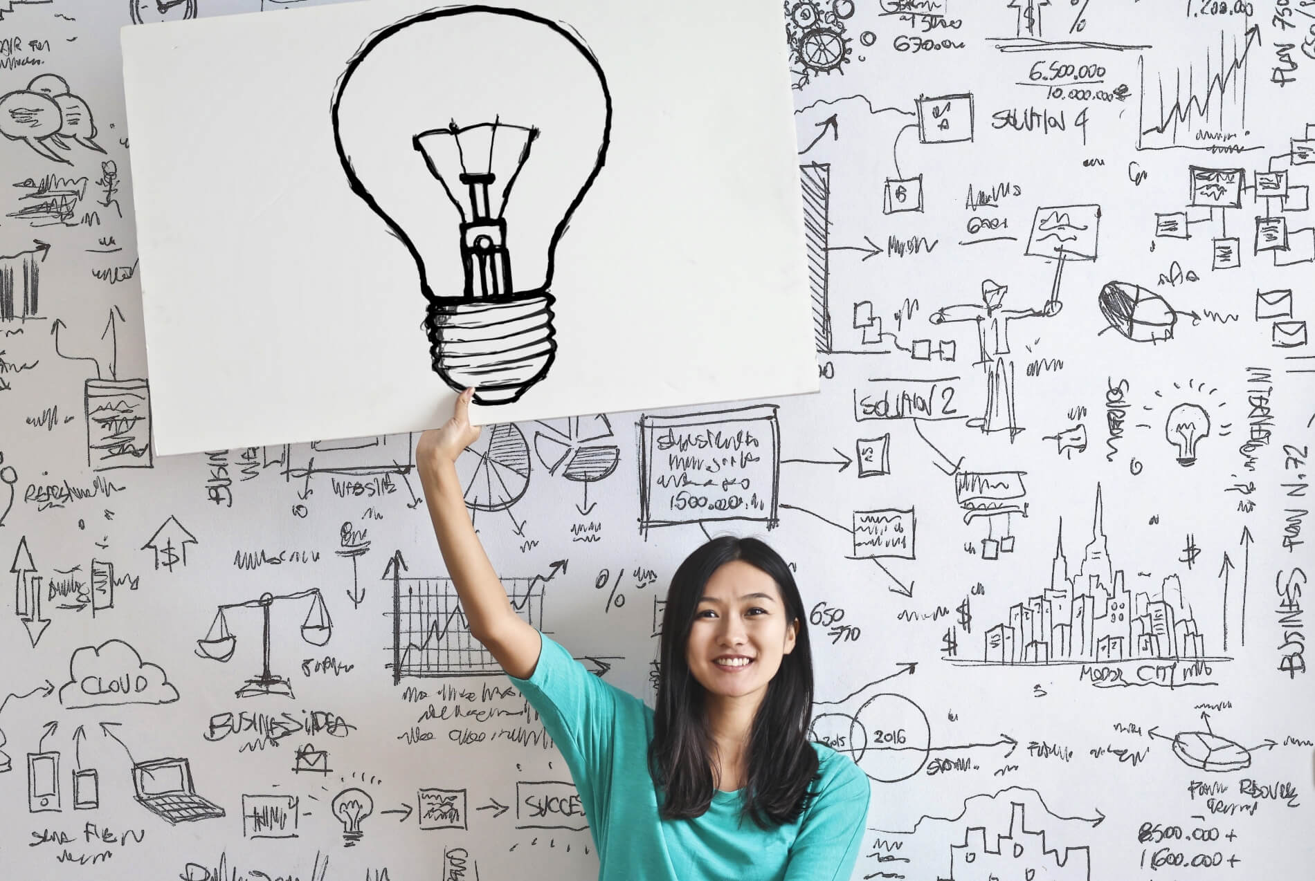 Woman holding a light-bulb sketch in front of a whiteboard full of other sketches and images.