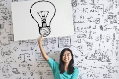 Woman holding a light-bulb sketch in front of a whiteboard full of other sketches and images.