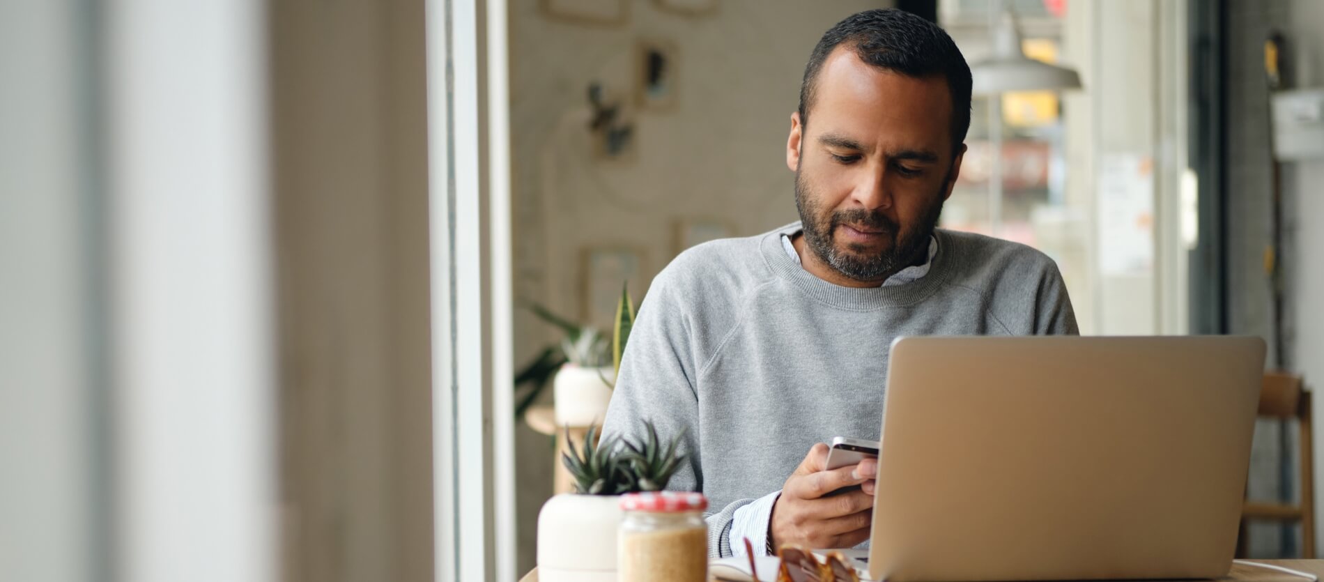 A man sitting by a window at a table using his laptop and phone